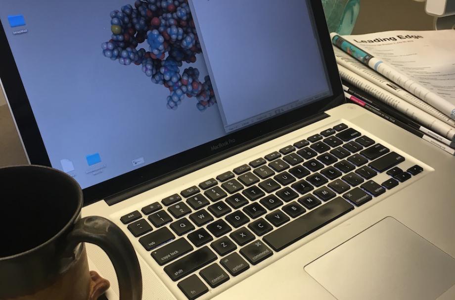 A laptop computer with the screen open. A coffee cup to the left of the computer and a stack of scientific magazines to the right.